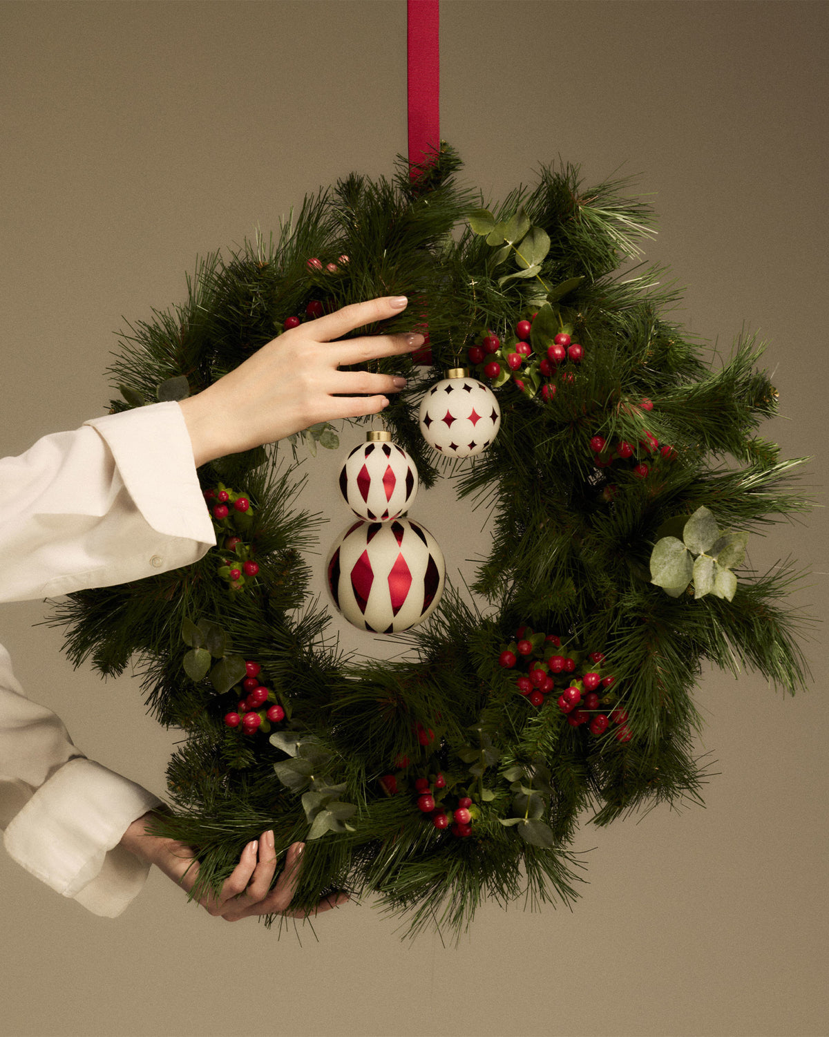 Two diamond-patterned and one star-patterned white and red Christmas ornaments by Alessi, held on a pine wreath with berries.