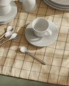 KU white cup by Alessi with a saucer, on a beige grid tablecloth alongside silver spoons.