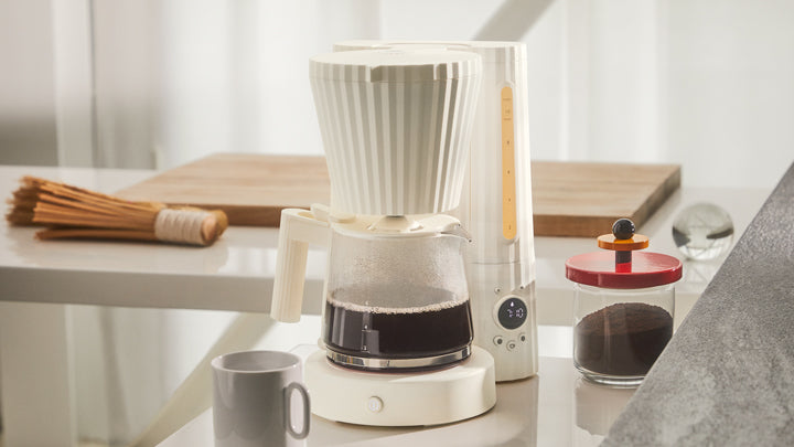 White drip coffee maker with glass carafe filled with dark coffee, digital timer, and red-lidded coffee jar beside it on a bright kitchen counter.
