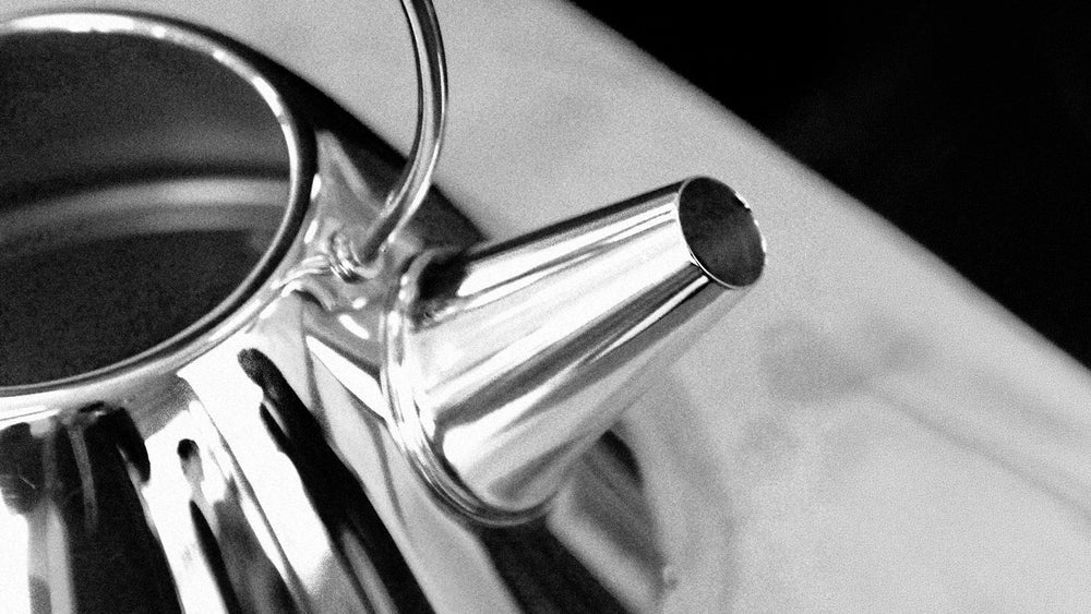 Shiny metallic kettle with long spout and circular handle, resting on light surface, black-and-white high-contrast photo.