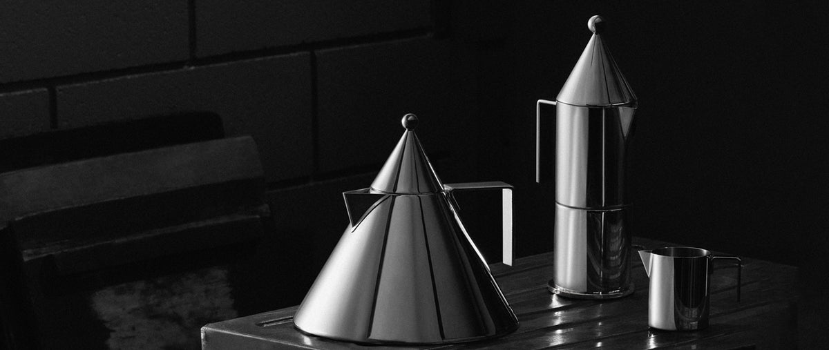 Shiny metallic cone-shaped kettle and matching pitcher on dark wooden surface, black brick background, monochrome lighting.