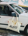 Cream canvas XL tote bag by Alessi featuring black handwritten 'ALESSI' text, hanging on a white car in a snowy mountain landscape.