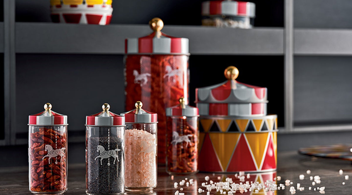 Colorful circus-themed glass jars with red and white striped lids, filled with spices and grains, arranged on a dark countertop with scattered salt crystals.