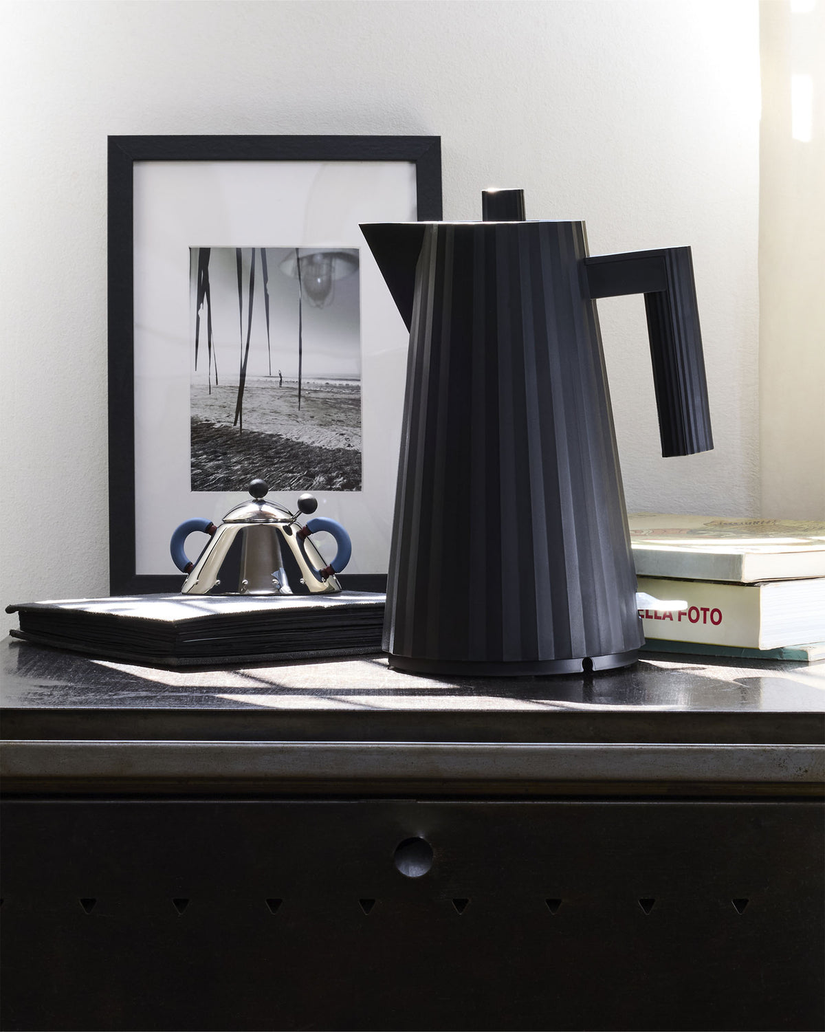 Black electric kettle by Alessi with a distinct pleated texture, styled on a dark dresser with a framed photo and books.