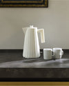 White pleated electric kettle by Alessi, standing on a dark countertop with two white mugs.