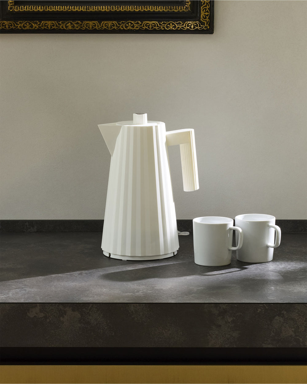 White pleated electric kettle by Alessi, standing on a dark countertop with two white mugs.