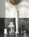 White ribbed hand blender by Alessi, with matching chopper, whisk attachment, and measuring cup on a dark counter.