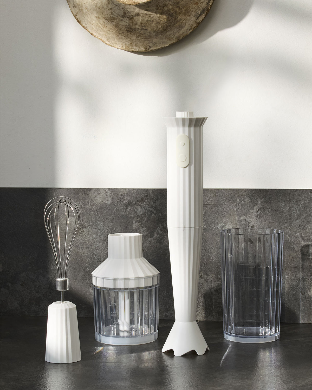 White ribbed hand blender by Alessi, with matching chopper, whisk attachment, and measuring cup on a dark counter.