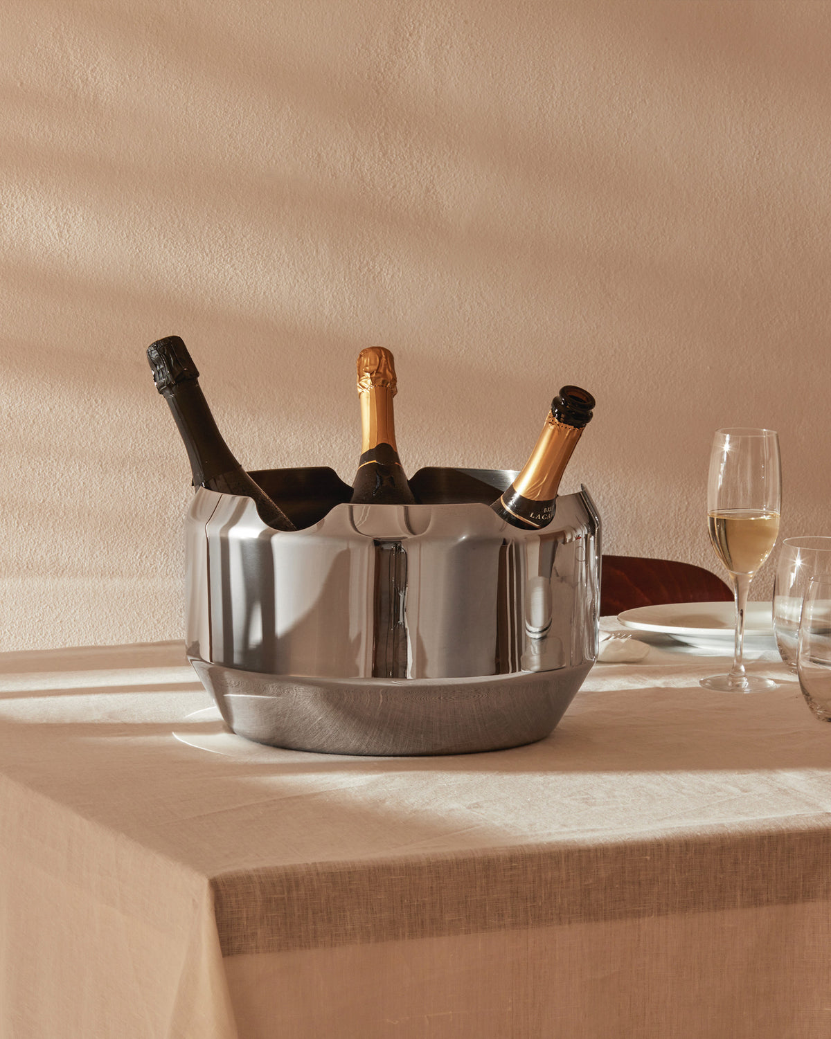 Silver segmented stainless steel champagne cooler by Alessi, holding three bottles on a linen tablecloth.