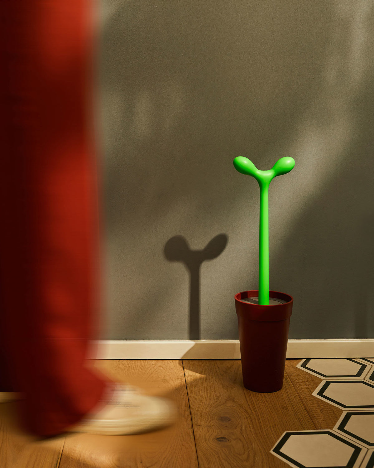 Green sprout-shaped toilet brush with red pot holder by Alessi, on a wooden floor against a grey wall.