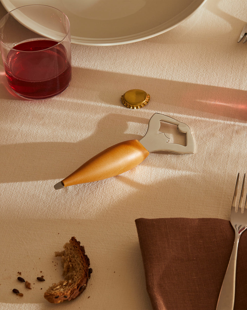 Wooden handle and silver metal bottle opener by Alessi, resting on a beige tablecloth next to a golden bottle cap.