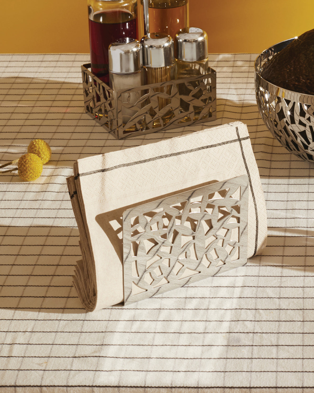 Silver-toned CACTUS! napkin holder by Alessi with abstract cutout design, alongside matching cruet set holder and bowl on a striped tablecloth.