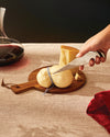 Silver Antechinus cheese knife by Alessi, cutting into soft yellow cheese on a wooden board.