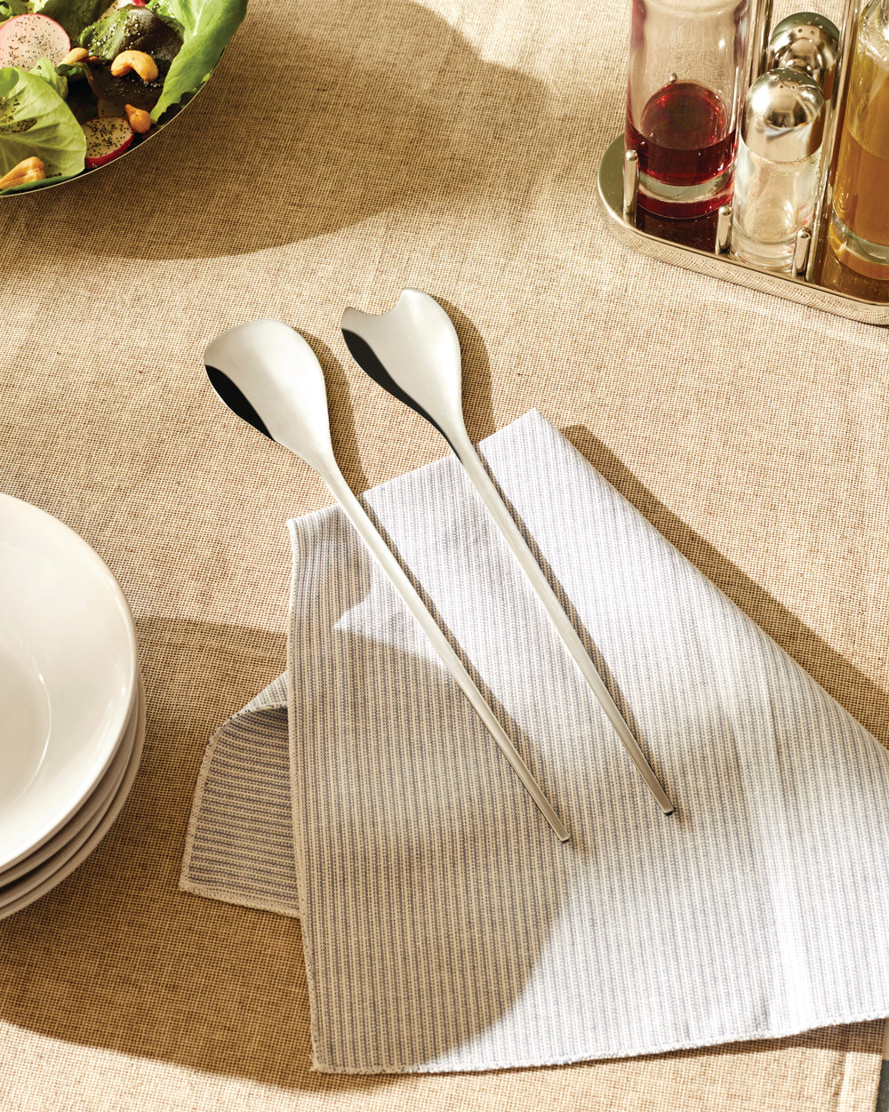 Sleek silver-toned salad servers by Alessi resting on a striped white and blue napkin.