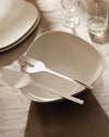 White serving bowl and matching utensils by Alessi, featuring an organic shape, dramatically lit on a linen tablecloth.