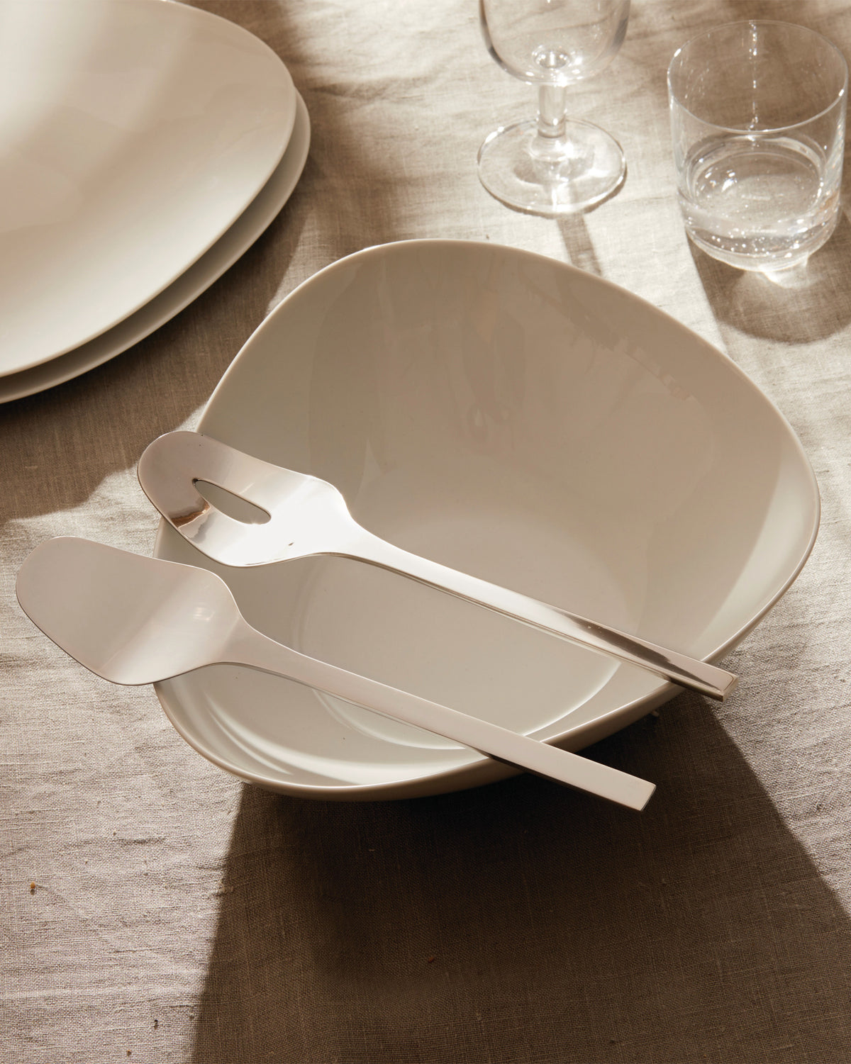 White serving bowl and matching utensils by Alessi, featuring an organic shape, dramatically lit on a linen tablecloth.