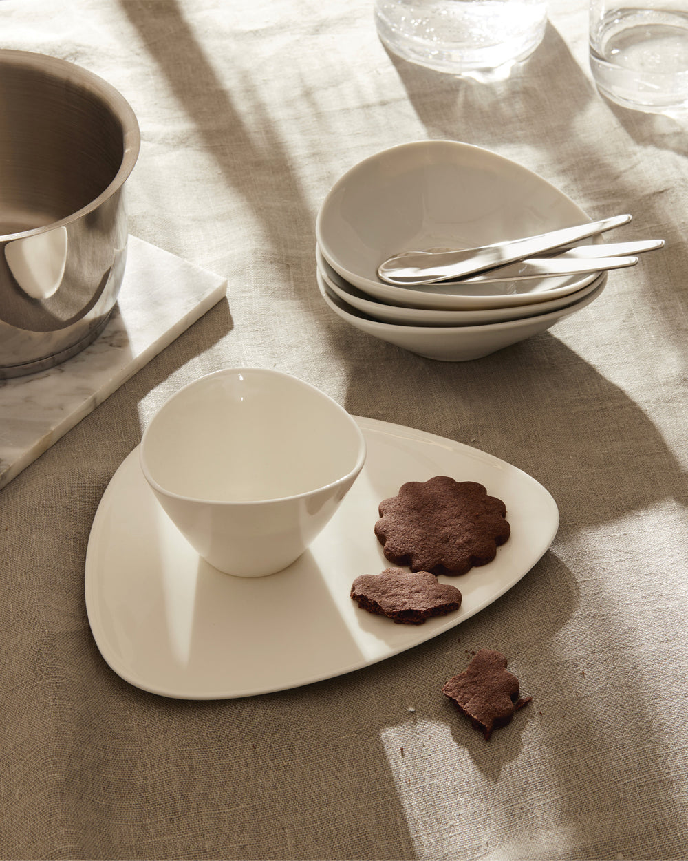 Irregularly shaped white porcelain bowl and platter set by Alessi, displaying chocolate cookies on a light linen tablecloth.