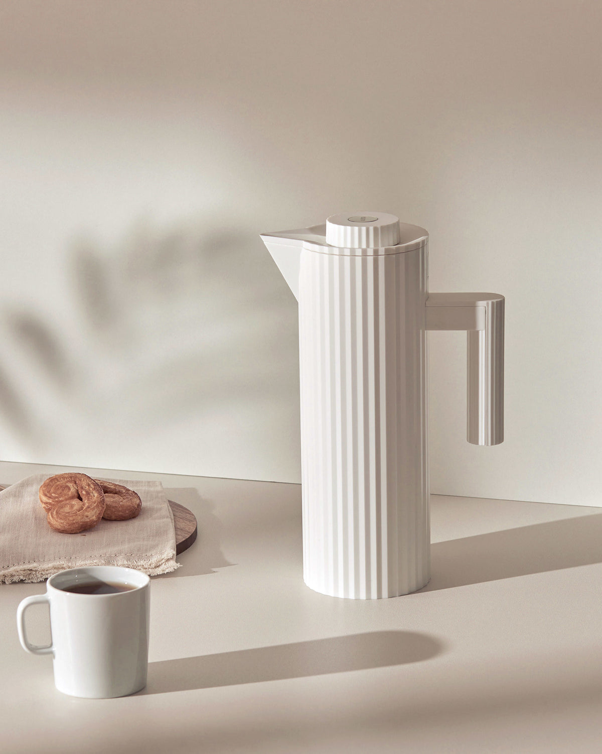 White pleated thermal carafe by Alessi with a sleek handle, next to a coffee mug and palmiers.