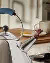 Shiny stainless steel whistle kettle by Alessi with a blue handle, black lid knob, and a red bird-shaped whistle.