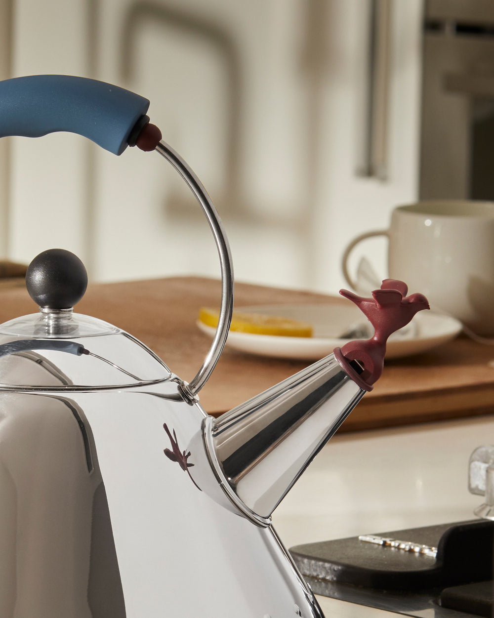 Shiny stainless steel whistle kettle by Alessi with a blue handle, black lid knob, and a red bird-shaped whistle.
