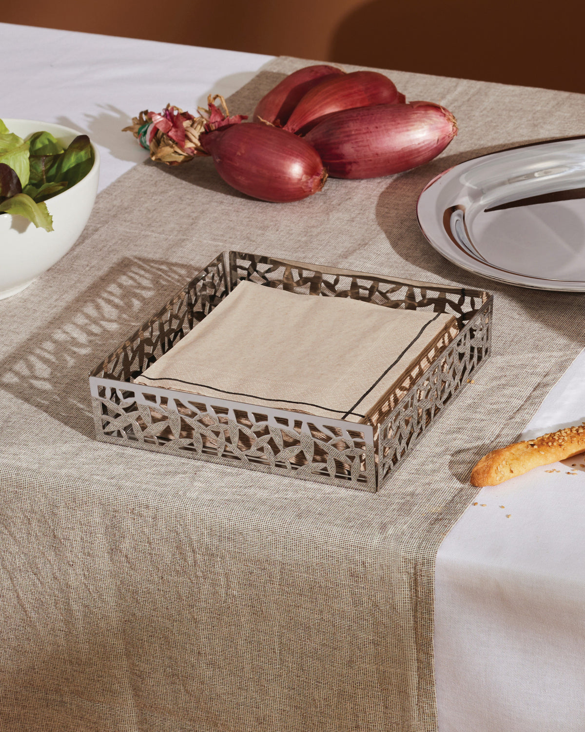 Silver metal napkin holder by Alessi with a geometric cactus cut-out design, holding beige napkins on a linen table runner.