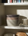 Stacked Tonale ceramic bowls in muted grey, pink, and taupe by Alessi, with a matching pitcher and wooden board.