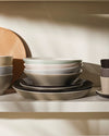 Stacked ceramic dinnerware by Alessi, in neutral and pastel shades, on a shelf with a wooden board.