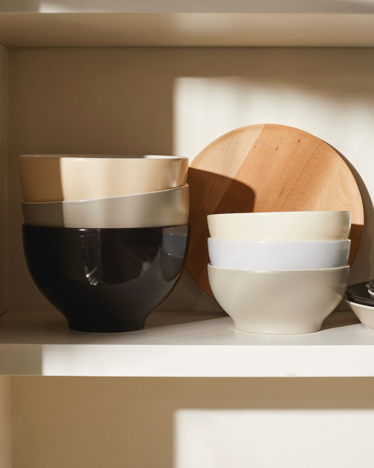 Ceramic bowls by Alessi, stacked in neutral colors on a shelf with a round wooden board.