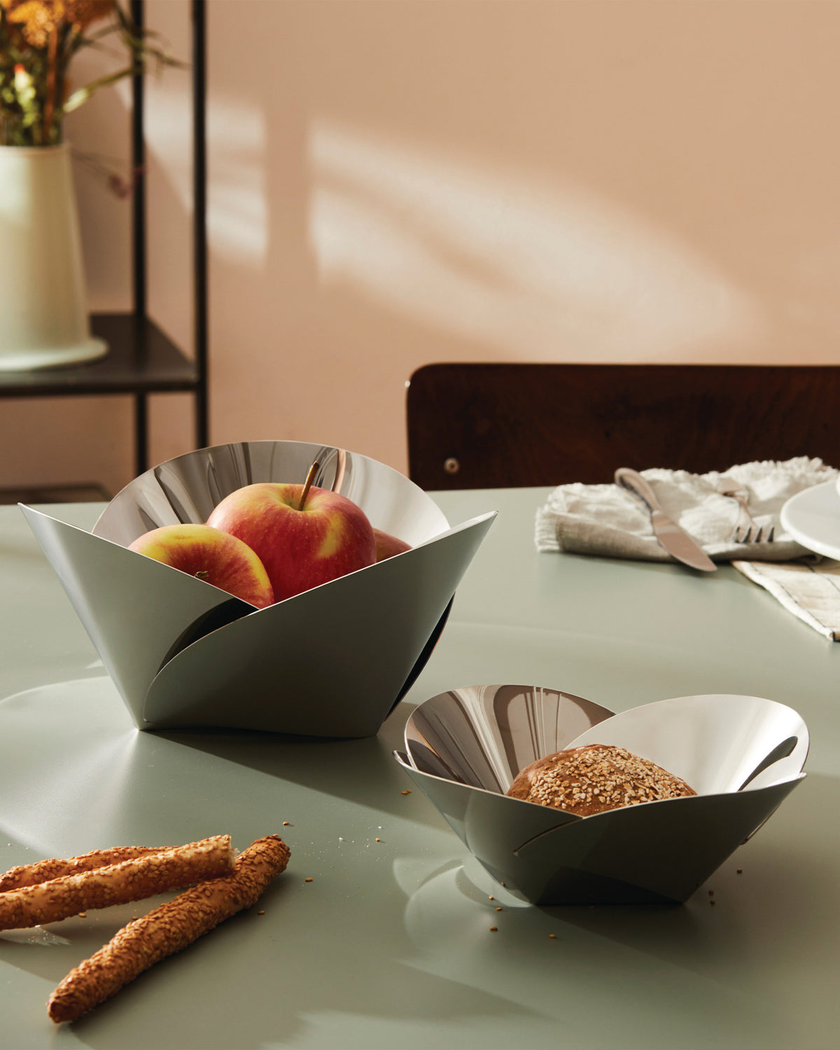 Geometric grey and shiny metal baskets by Alessi, holding fruit and bread.