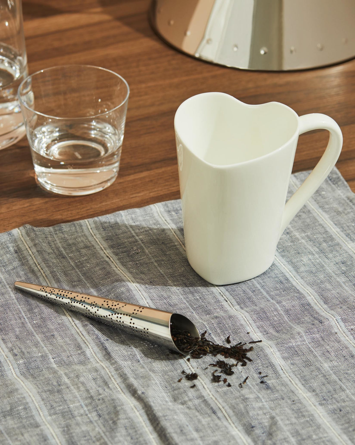 White heart-shaped ceramic mug by Alessi, on a striped blue napkin next to a metal tea infuser and loose tea.
