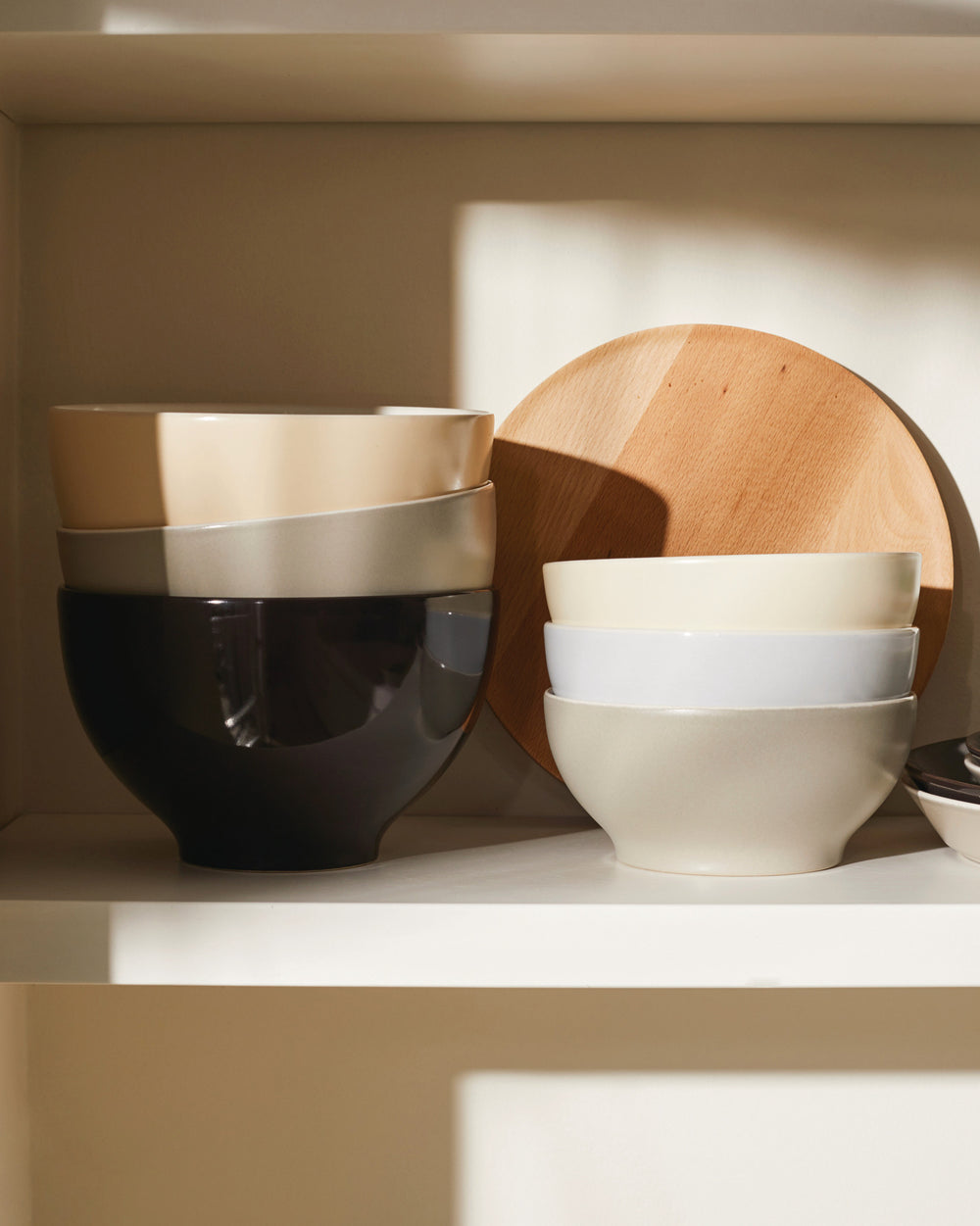 Stacked ceramic bowls in neutral colors by Alessi, on a white shelf next to a wooden board.