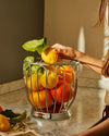 Polished stainless steel fruit basket by Alessi, filled with lemons and oranges on a marble counter, a hand reaches inside.