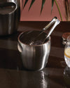 Stainless steel ice bucket with tongs by Alessi, containing ice cubes and showing condensation, next to a glass with lemon on a dark table.