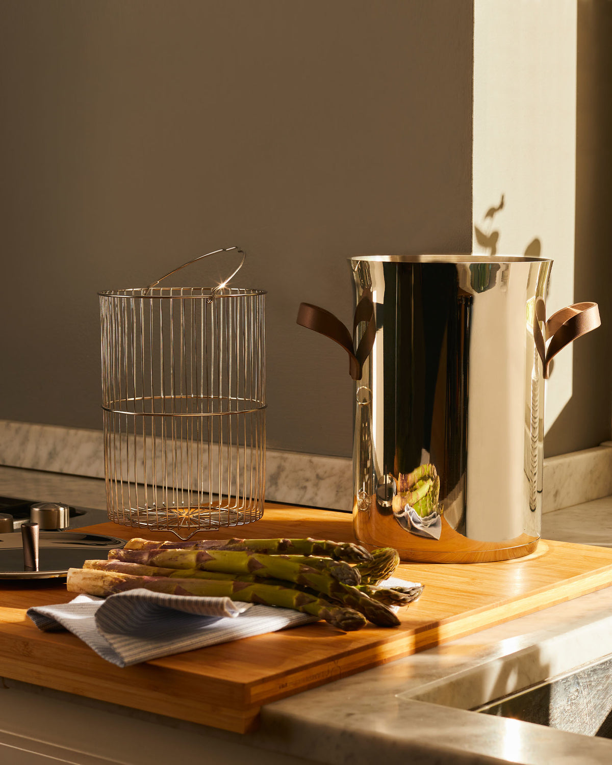 Tall stainless steel pot by Alessi with brown leather handles, alongside a gold wire insert basket and fresh asparagus.