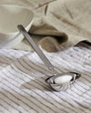Silver stainless steel ladle by Alessi with a brushed handle, resting on a striped linen napkin next to a white bowl.