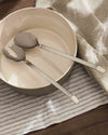 Silver-toned serving set by Alessi, placed in a cream ceramic bowl on a striped linen cloth.