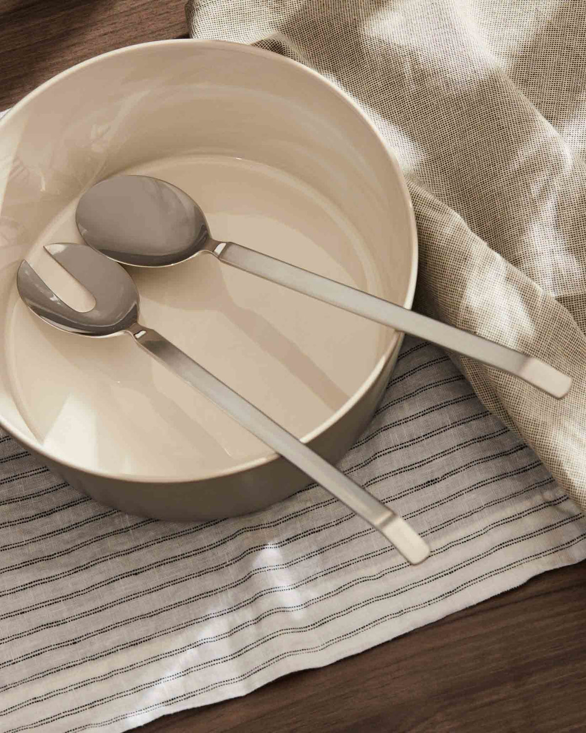 Silver-toned serving set by Alessi, placed in a cream ceramic bowl on a striped linen cloth.