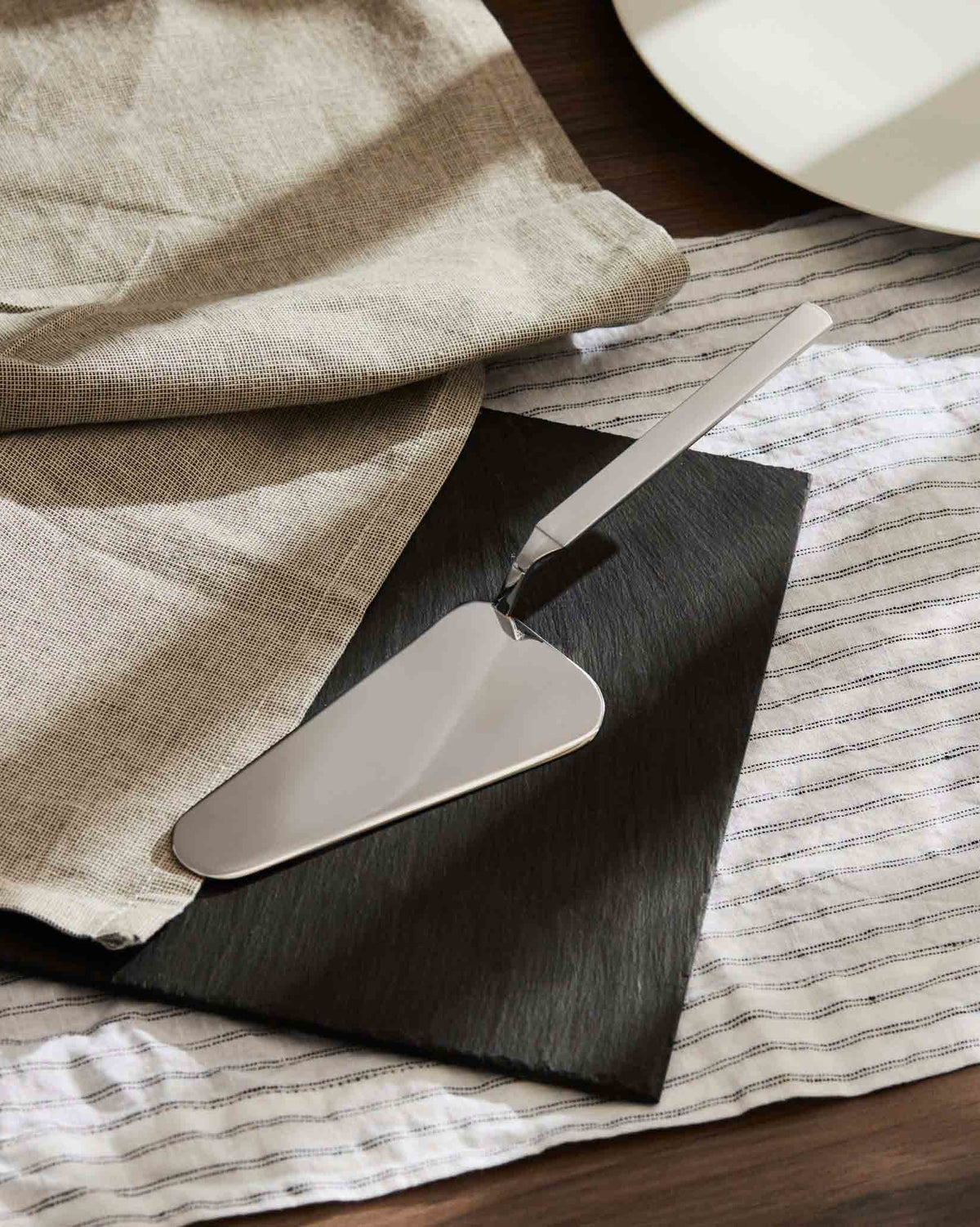 Shiny silver cake server by Alessi, resting on a black slate board, with a beige linen napkin and striped white cloth.