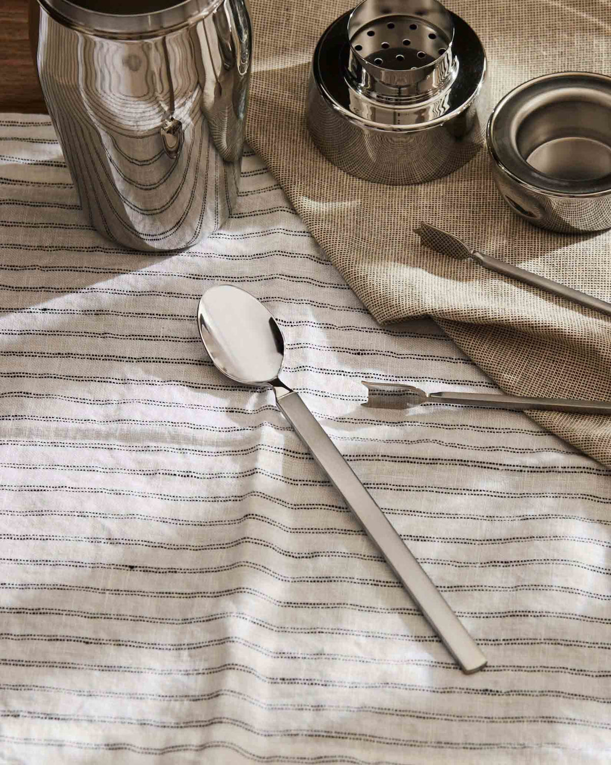 Stainless steel spoon by Alessi with a polished bowl and matte handle, on a striped white linen cloth.