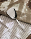 Silver metal scoop by Alessi resting on beige and striped white linen fabrics.