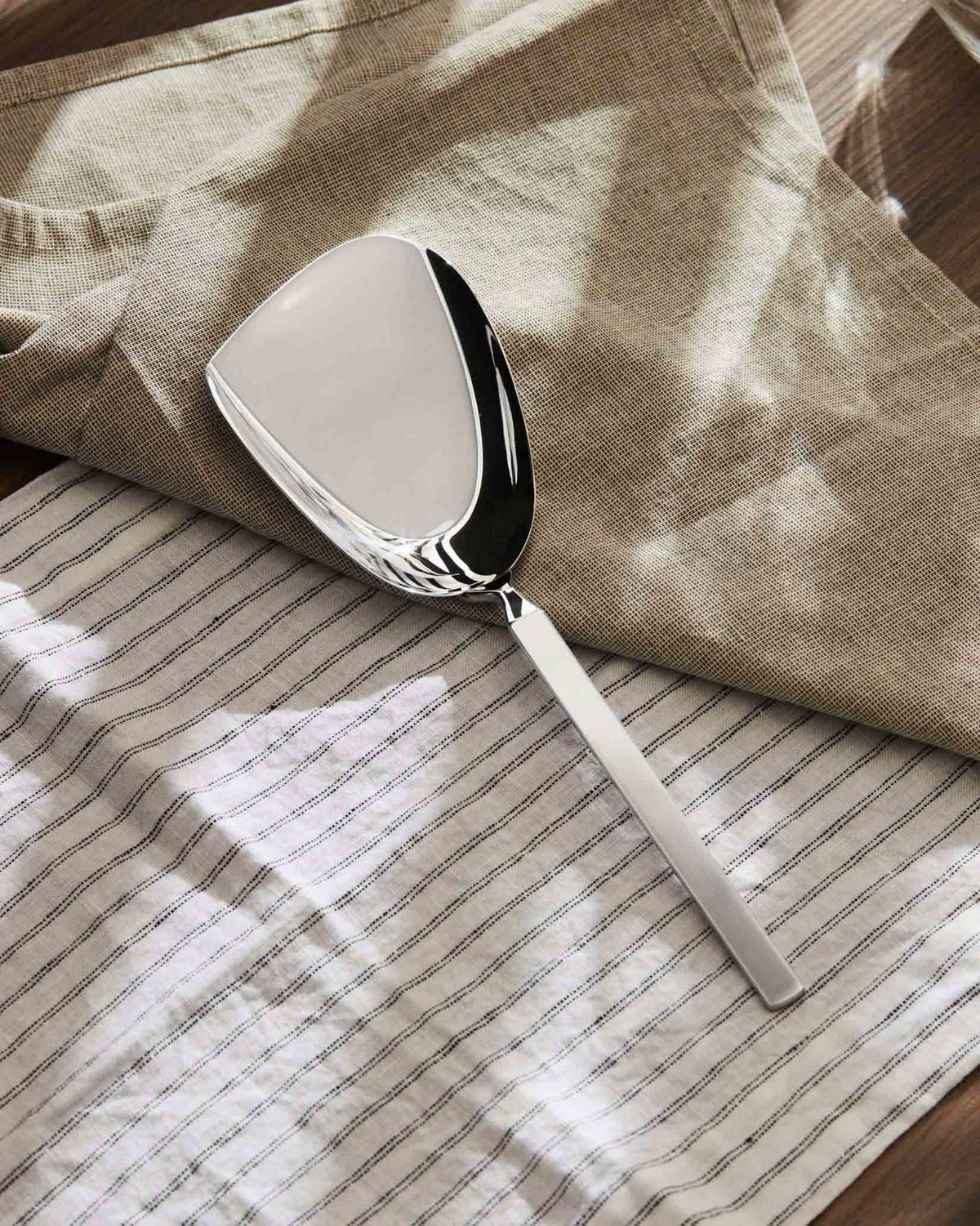 Silver metal scoop by Alessi resting on beige and striped white linen fabrics.