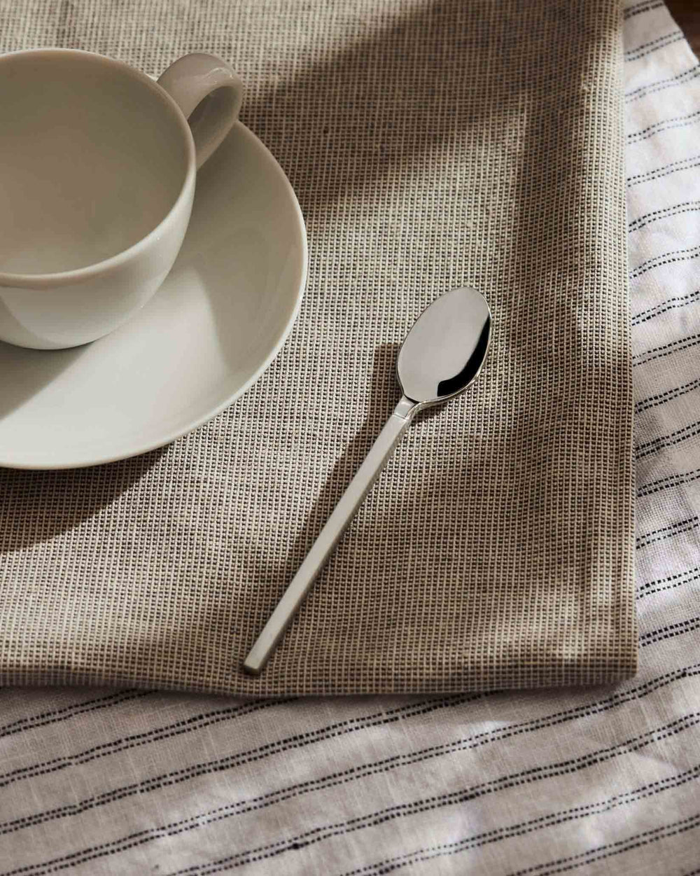 Polished silver spoon by Alessi with a sleek, faceted handle, shown on a woven placemat with a white cup.