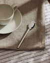 Silver metal spoon by Alessi resting on a beige woven placemat with a white striped cloth and ceramic cup.