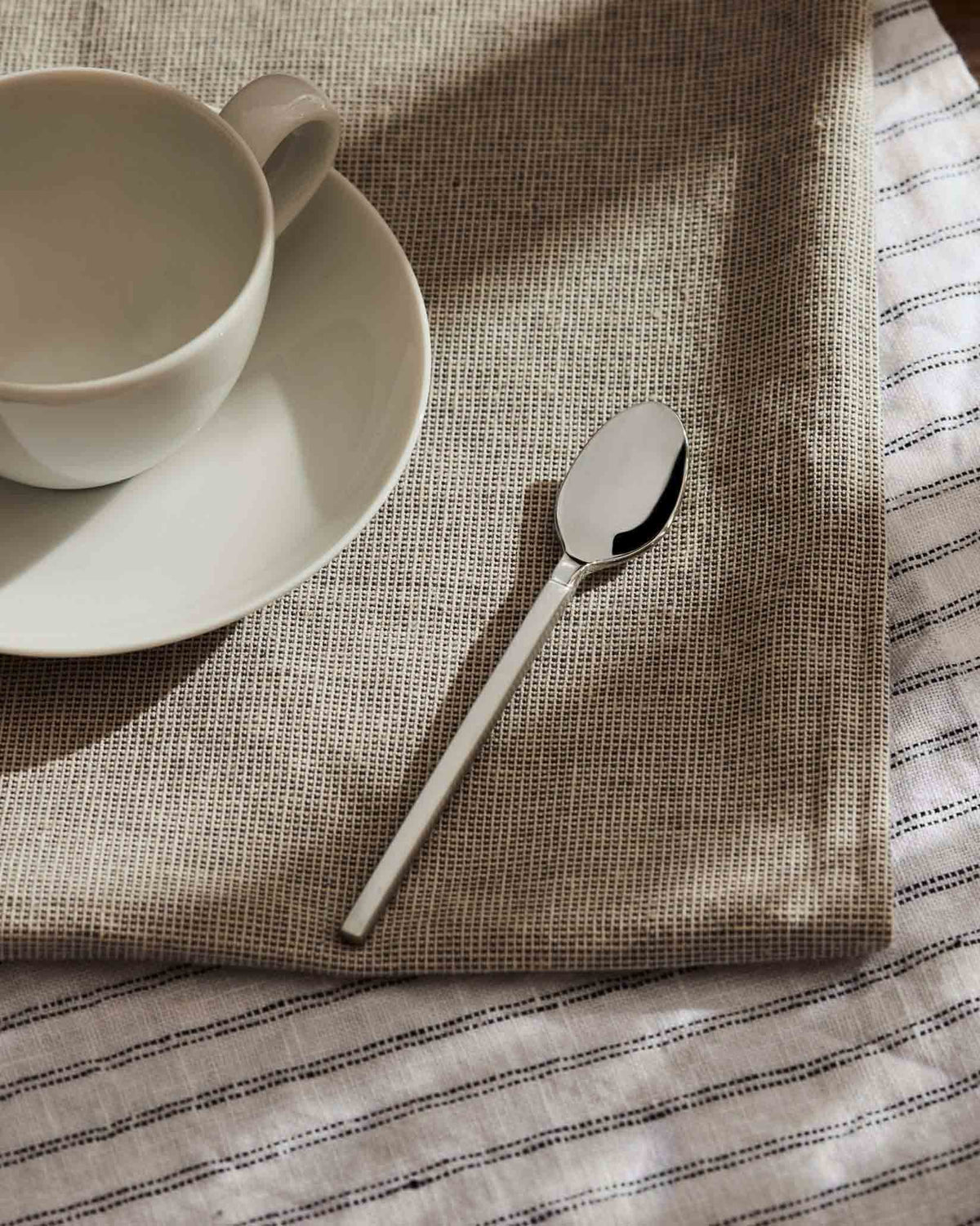 Silver metal spoon by Alessi resting on a beige woven placemat with a white striped cloth and ceramic cup.