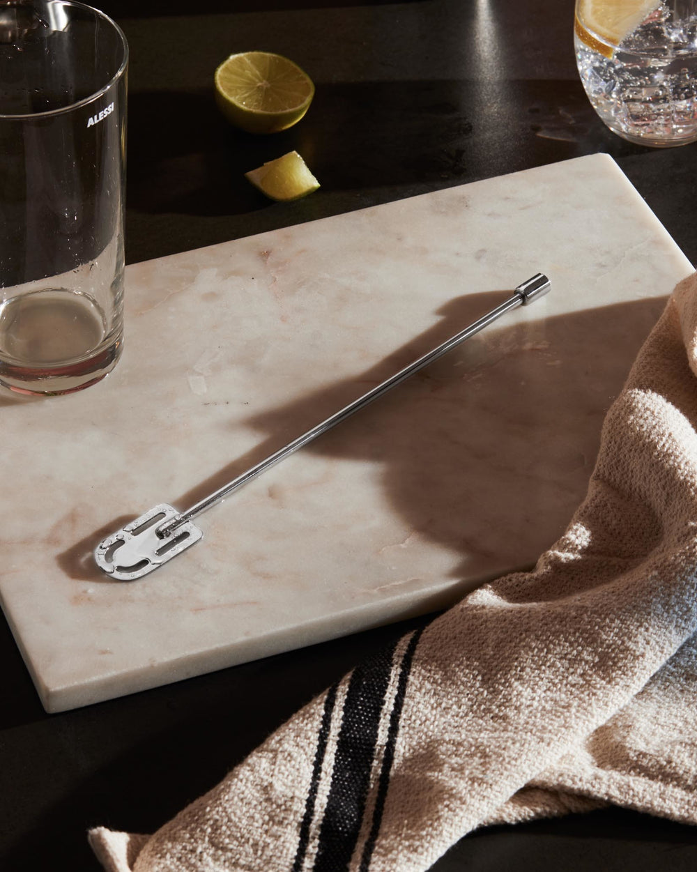 Silver metal cocktail stirrer by Alessi with a slotted end, on a marble board next to a glass, lime, and beige striped towel.