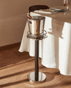 Polished stainless steel champagne bucket on a dark stand by Alessi, holding a bottle next to a dining table on a wooden floor.