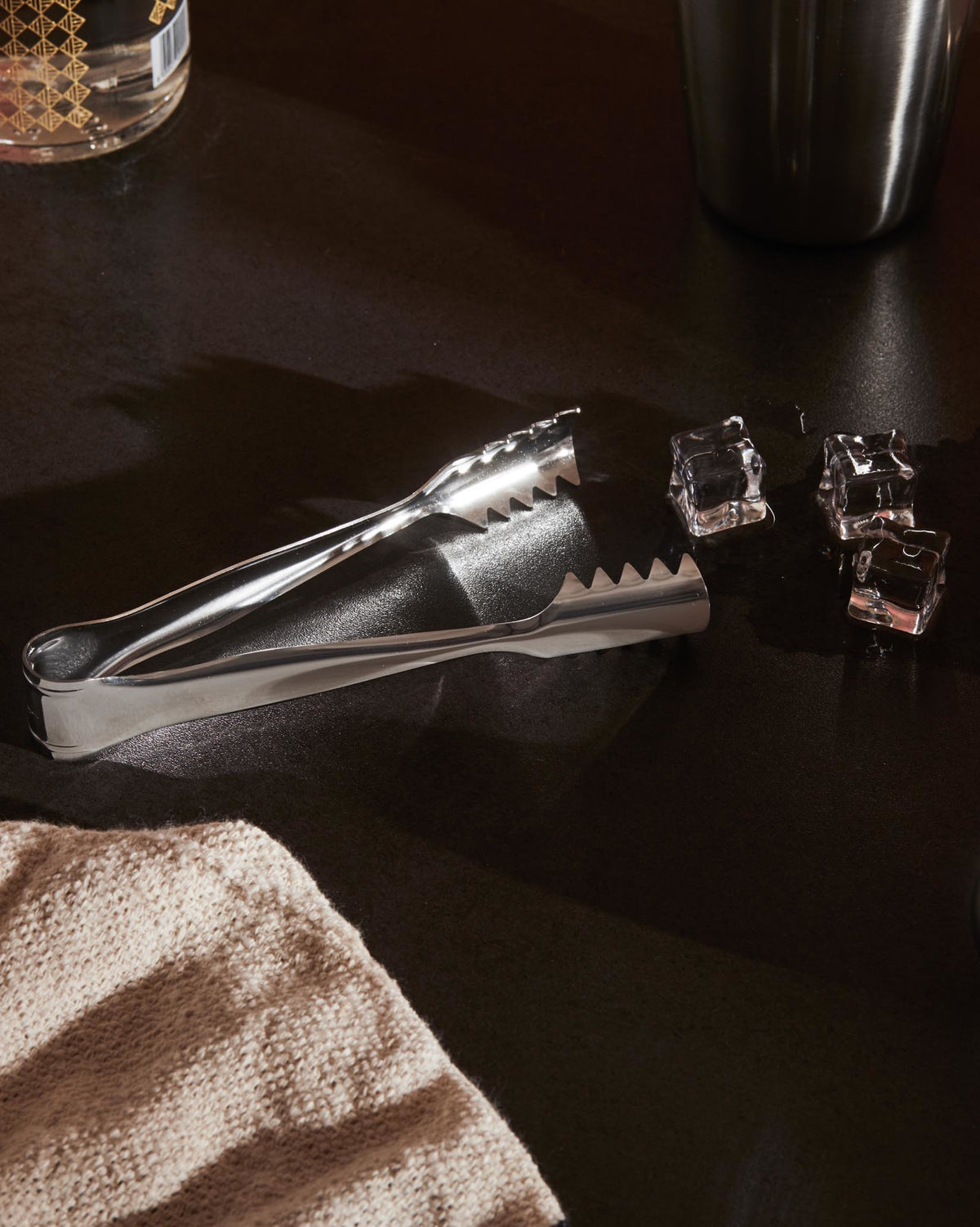 Stainless steel ice tongs by Alessi with serrated edges, next to ice cubes on a dark surface.