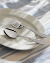 Polished silver flatware set by Alessi, a spoon and fork on a white plate over a blue and beige striped tablecloth with shadows.