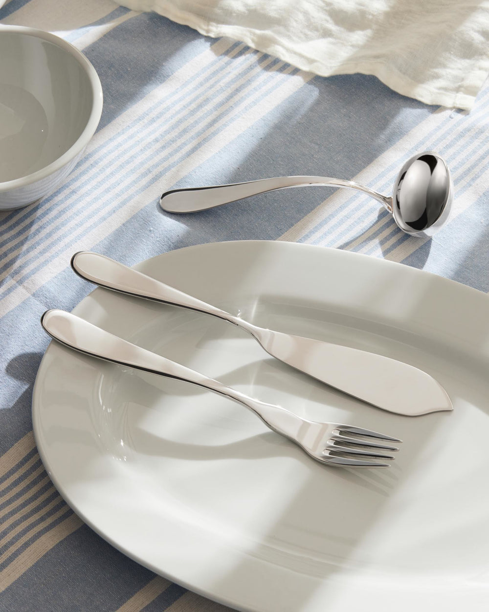 Silver cutlery set by Alessi, sleek design on a white platter with a matching ladle on a blue and white striped tablecloth.