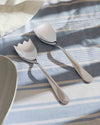 Silver salad servers by Alessi with a contemporary design, one featuring a forked edge, on a blue and white striped tablecloth, next to a white bowl.